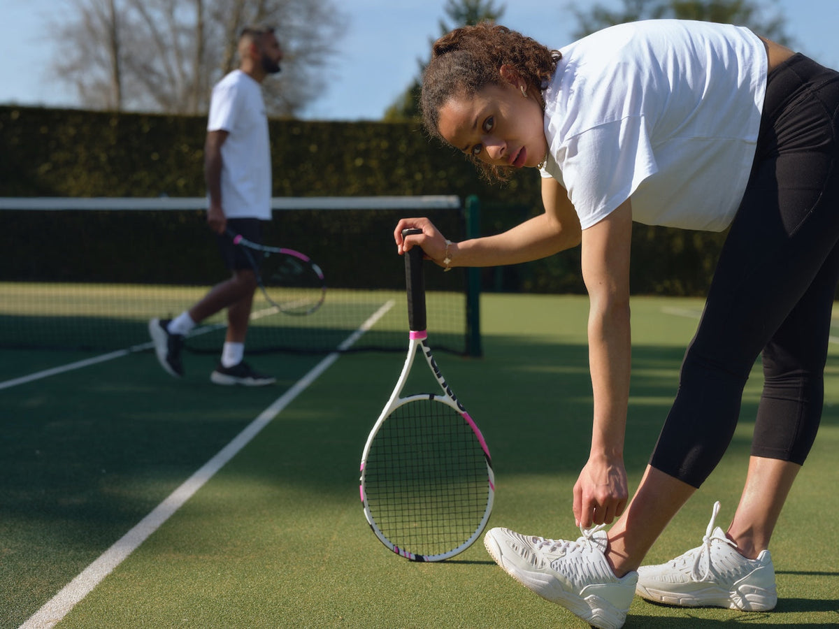 Woman in white t-shirt and black leggings tying white tennis shoes on a green court with racket.