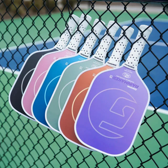 A colorful row of Gamma pickleball paddles leaning against a chain-link fence on a court.