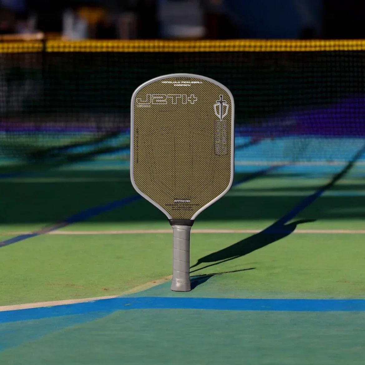 A professional J2Ti+ pickleball paddle standing upright on a blue and green court.