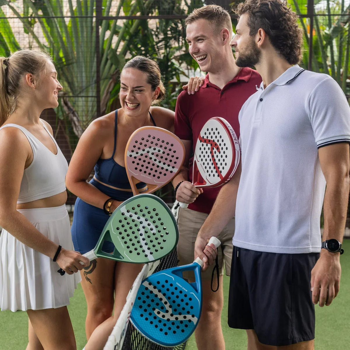 Four friends laughing and holding colorful padel rackets on an outdoor court.